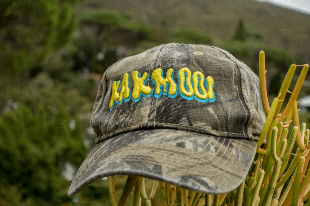 Faded olive green camo cap displaying the word "KAKMOOI" in a slimey font in 3D embroidery. Everyone loves a cool camouflage cap. New cap, funny cap, camper cap, washed cotton, Irony, Humor, meme. Hunting hat.